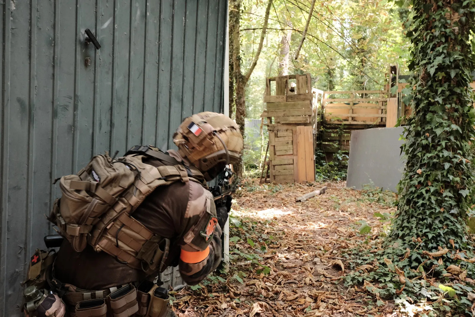 Joueur d'airsoft en tenue tactique, à couvert le long d'un bâtiment sur un terrain boisé.